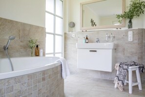 A modern bathroom with a freestanding bathtub, a vanity with a mirror, and a small stool with a blanket. - Ferienhaus, Fuhlendorf (Fuhlendorf)