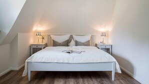 A bedroom with a sloped ceiling, a large bed with white bedding, two bedside tables with lamps, and a magazine on the bed. - Ferienhaus, Fuhlendorf (Fuhlendorf)