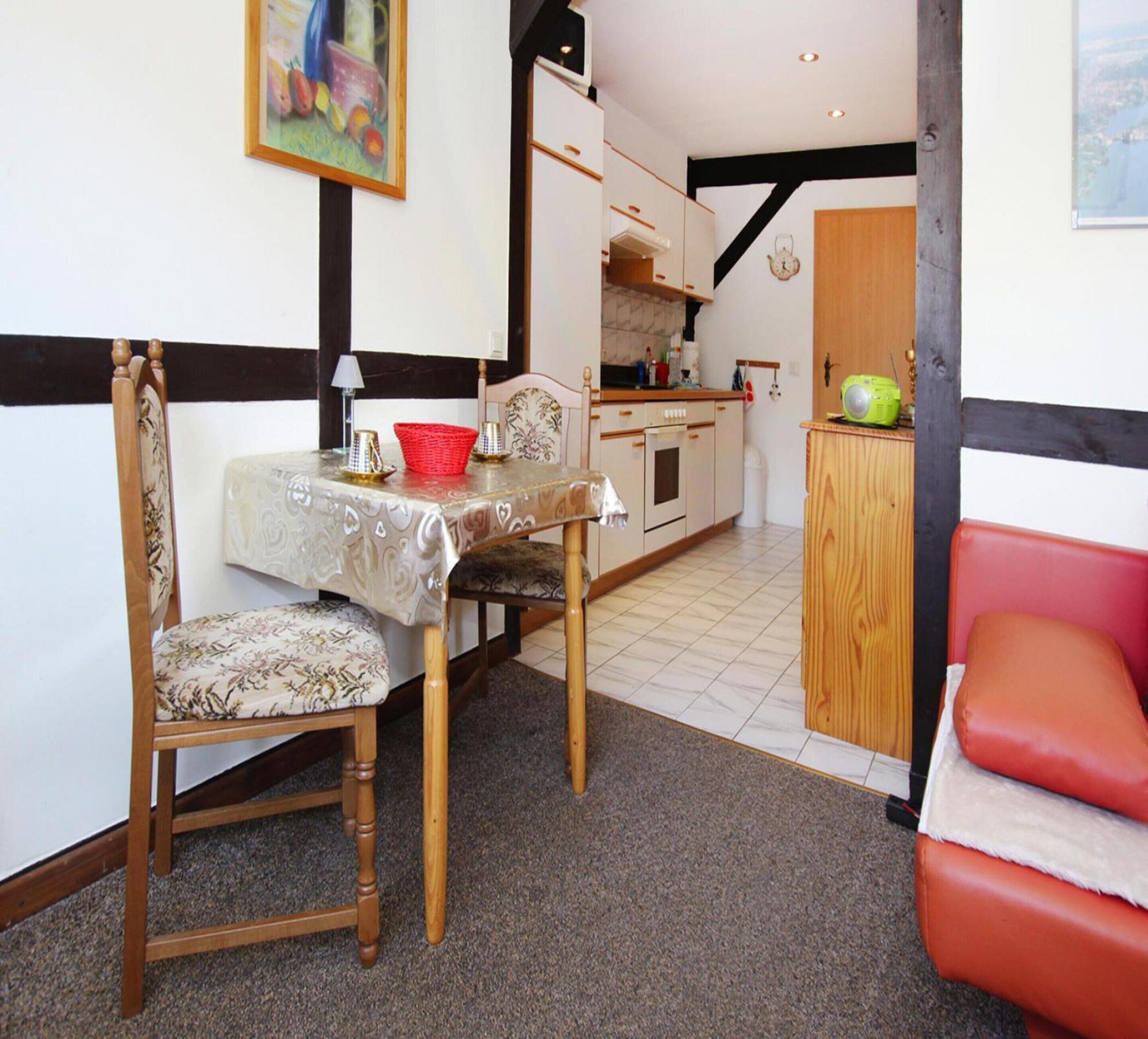A dining area with a table set for two, a kitchen in the background, and a red sofa.