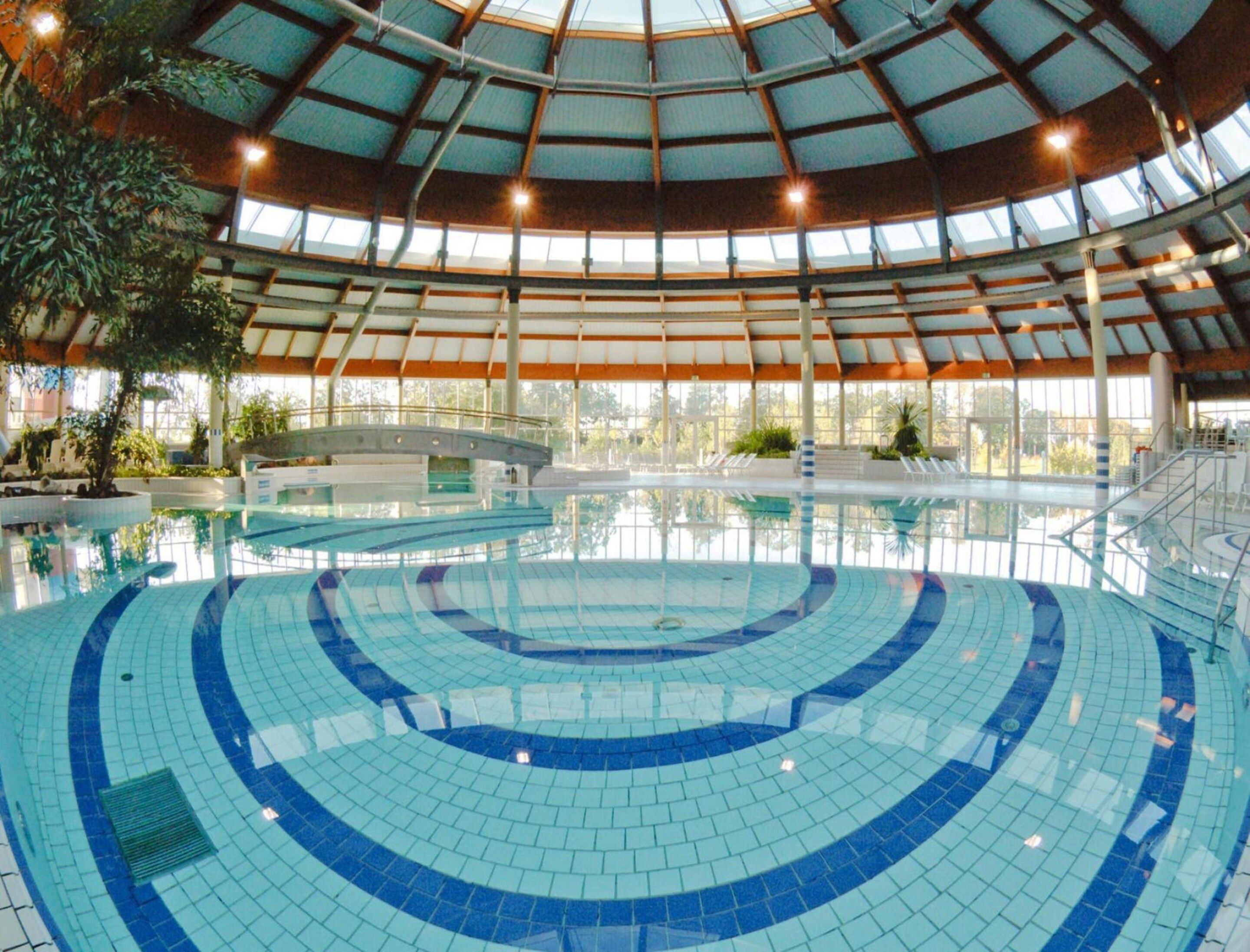 Uma piscina interior com design curvo, rodeada por uma área coberta com grandes janelas.