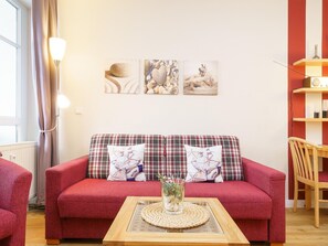 Um sofá vermelho com almofadas xadrez, uma mesa de centro de vidro com um jogo americano trançado e obras de arte na parede retratando cenas abstratas e paisagens.