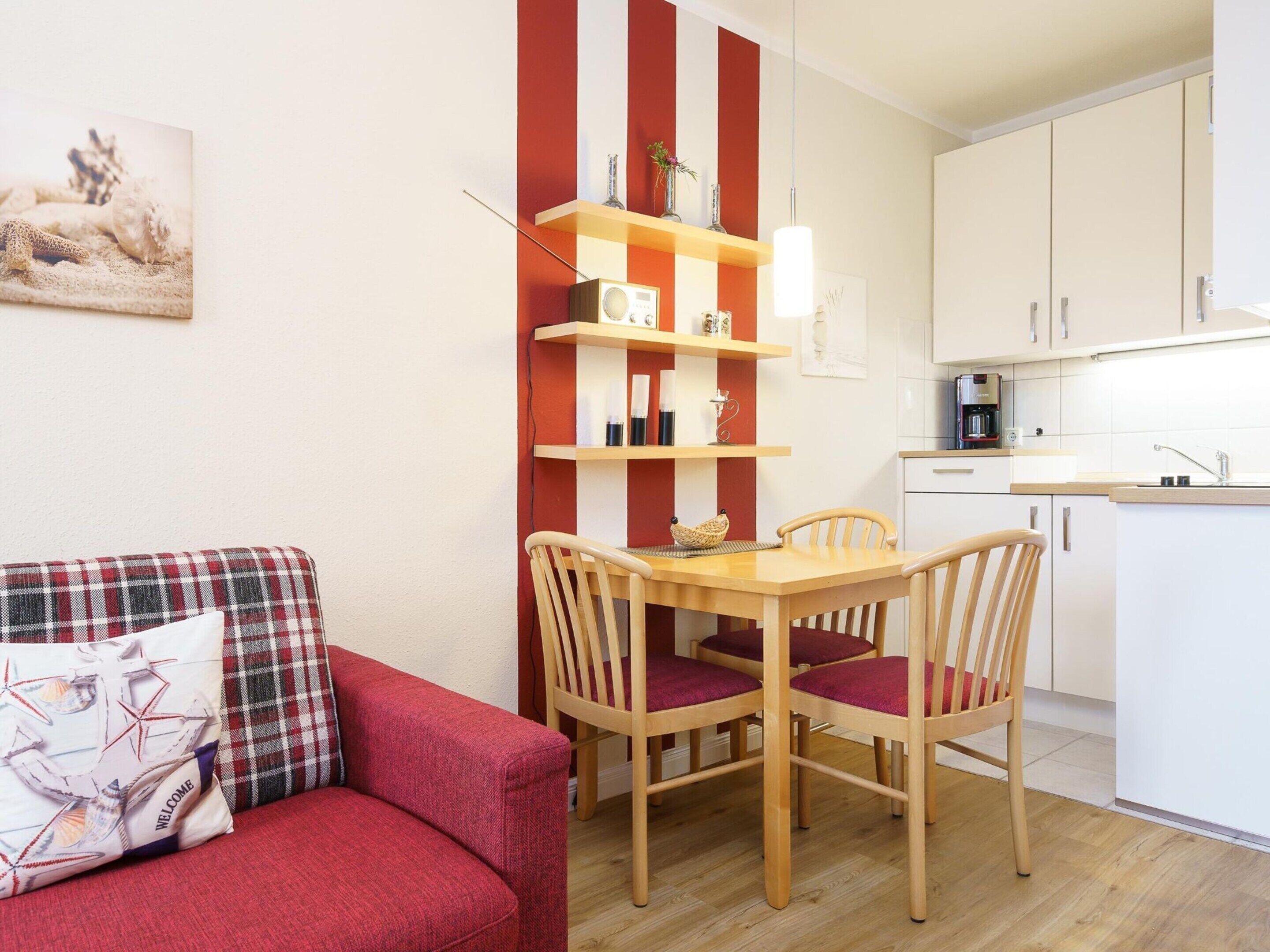 A compact living space with a red and white striped wall, a wooden dining table with four chairs, a red sofa with plaid cushions, and a kitchen area with white cabinets and a coffee machine.