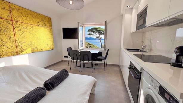 A modern hotel room with a bed, a dining area with a view, a kitchen with a washing machine, and a large yellow artwork on the wall. - Luxueux Haut de Villa 4 Pers à 30m de la Plage (Sainte-Maxime)