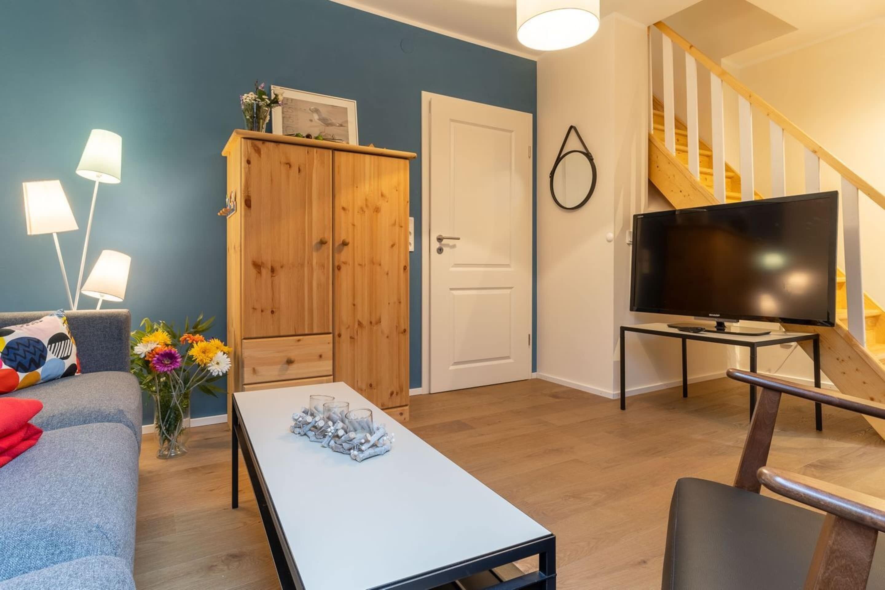 Una sala de estar con un sofá, una mesa baja con vasos, un armario de madera, un televisor sobre un mueble bajo y una escalera.