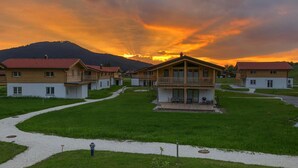 A row of wooden cabins with a central building featuring a balcony, surrounded by a grassy area and a mountain in the background. - Chalet Chiemgau, Inzell (Inzell)