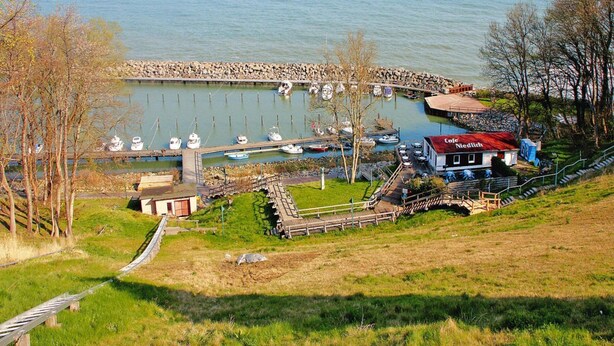 Un puerto deportivo con barcos amarrados, un pequeño edificio que dice 'Cafe Nedlich' y una zona verde con un sendero de madera.
