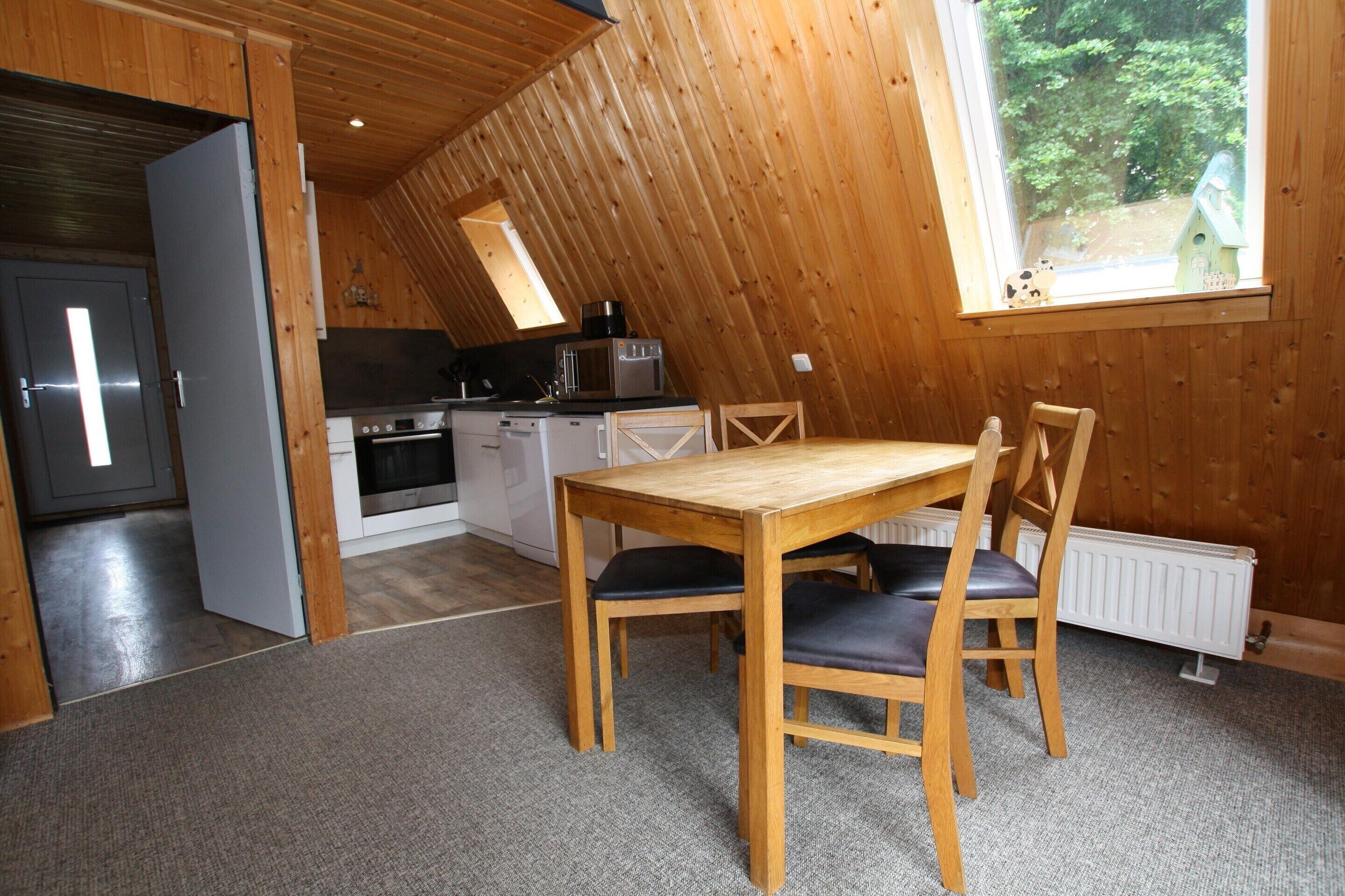 Um quarto de sótão acolhedor com teto de madeira, uma mesa de jantar com cadeiras, uma cozinha com eletrodomésticos e uma janela com vista para o verde.
