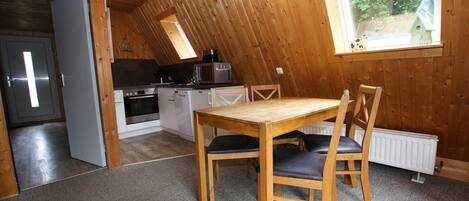 Um quarto de sótão acolhedor com teto de madeira, uma mesa de jantar com cadeiras, uma cozinha com eletrodomésticos e uma janela com vista para o verde.