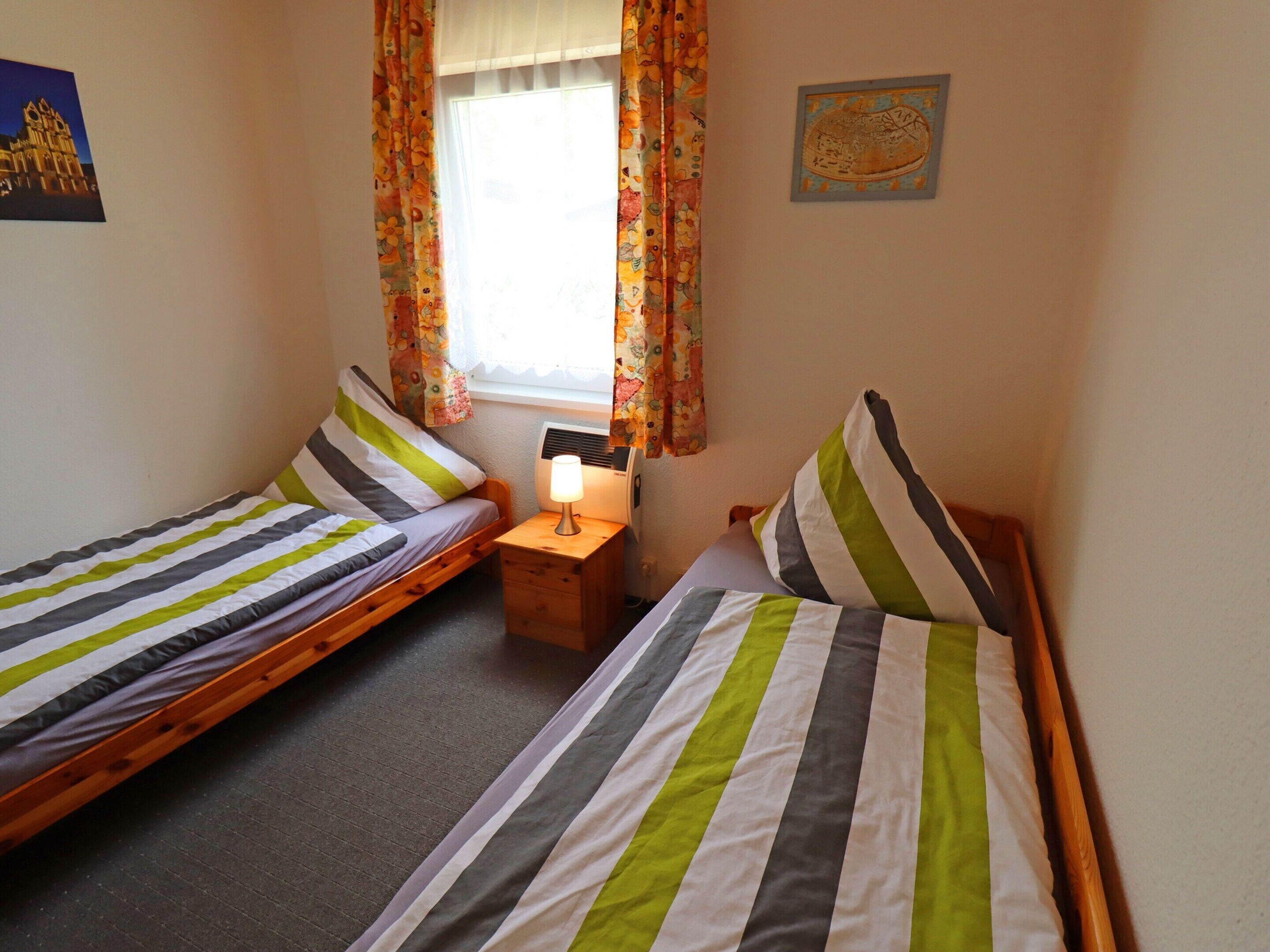 Una habitación con dos camas individuales, una mesita de noche con lámpara, una ventana con cortinas de flores y un cuadro colgado en la pared.