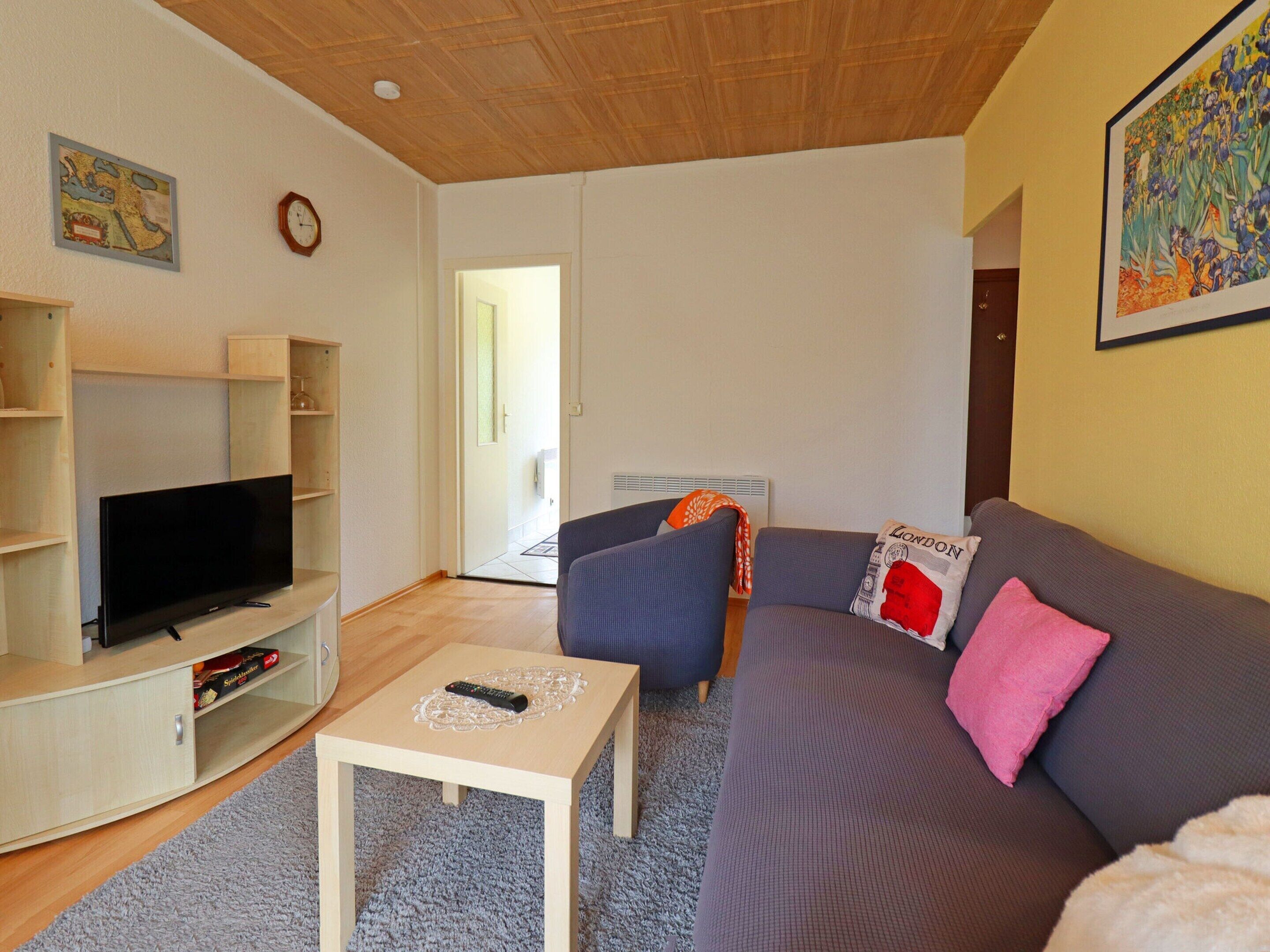 A living room with a flat-screen TV, a grey sofa with a pink pillow, a small white coffee table, and a blue armchair.