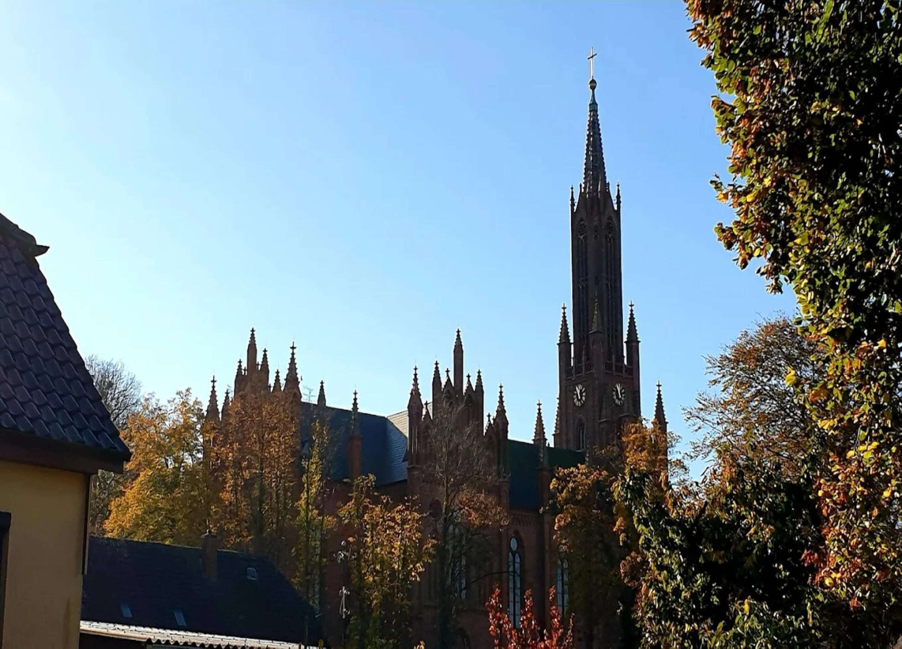 A church with a tall spire and multiple towers, surrounded by trees with autumn foliage.