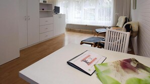 A modern hotel room with a wooden floor, white furniture, a desk with a chair, a radiator, and a window with curtains. - Ferienwohnung in Westerland auf Sylt (Sylt)