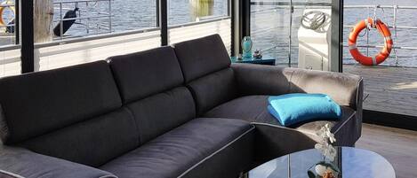 Een moderne zithoek met een donkergrijze bank, een glazen salontafel met decoratieve objecten, en uitzicht op een steiger en water door grote ramen.