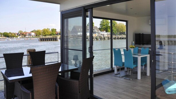 Ein Balkon mit Korbmöbeln und einem Tisch, mit Blick auf ein Ufer mit Gebäuden und einem Steg.