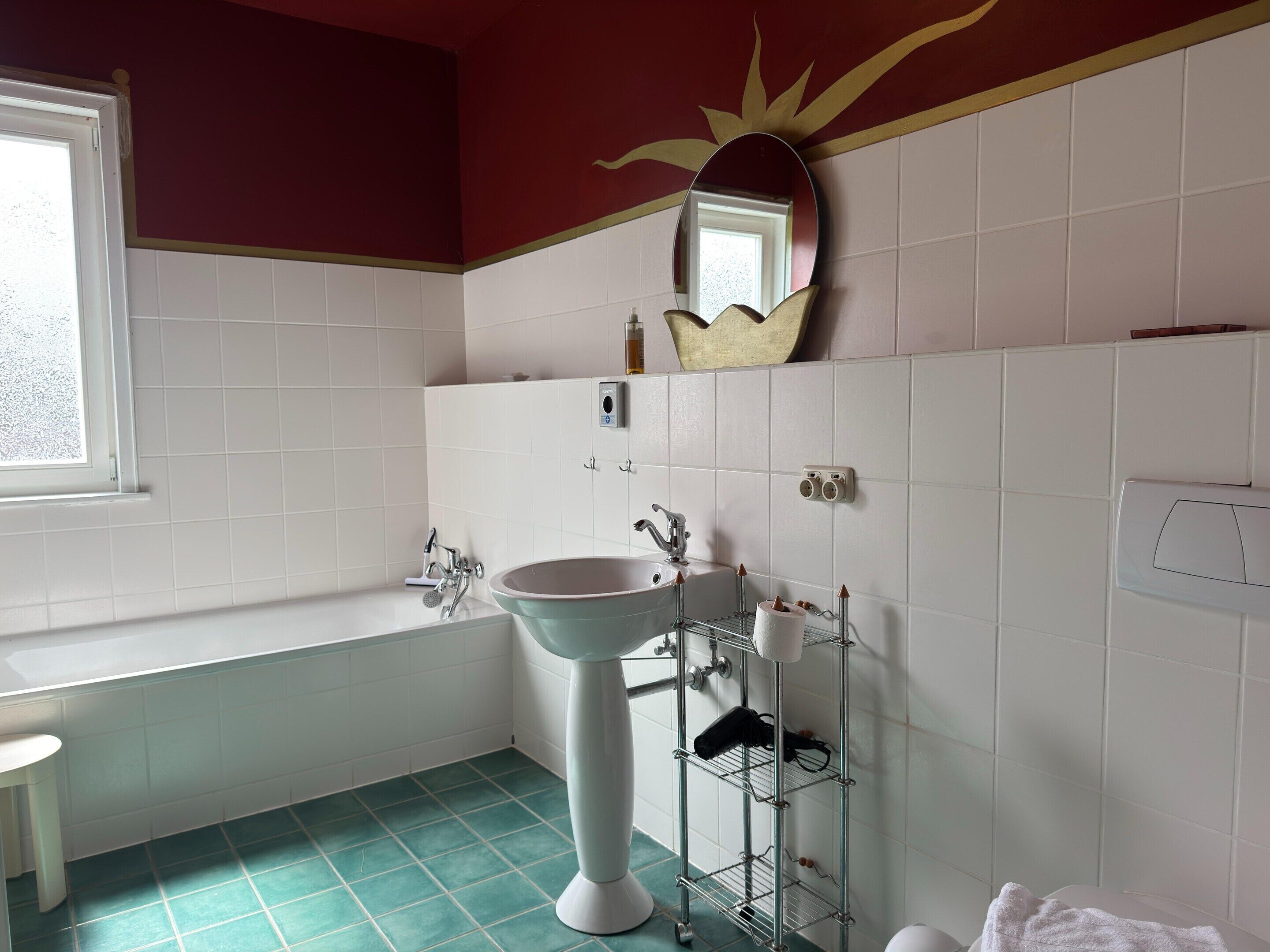 A bathroom with a pedestal sink, a bathtub, a mirror, and a shelf with toiletries.