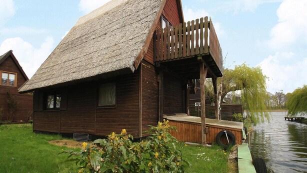 A wooden house with a thatched roof, a wooden porch, and a small dock extending into a body of water. - Bootshaus in Teterow - Formerly TUI (Teterow)