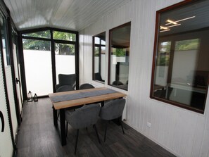 A room with a wooden table and four chairs, large windows with blinds, and a view of a white building outside.