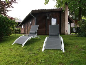 Two reclining chairs in a grassy area in front of a wooden house with a window and a door.