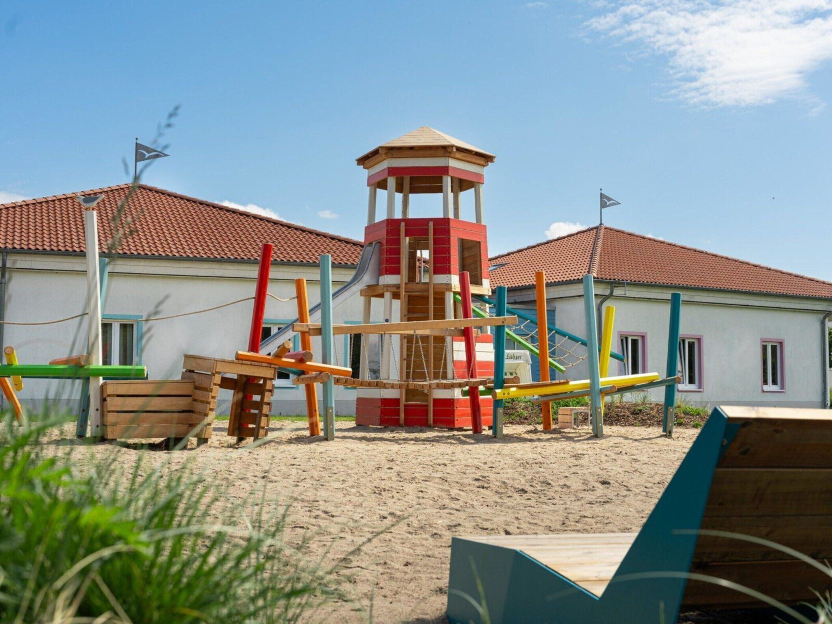 A playground with a climbing structure and slides, set against a building with a red-tiled roof.