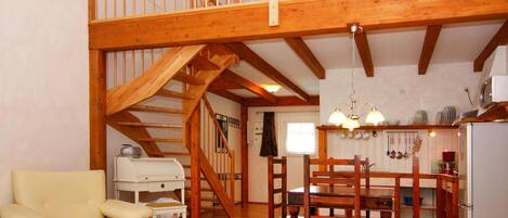 A spacious living area with a wooden staircase, a dining table with chairs, a sofa, and a kitchen area with a refrigerator and various kitchen items.