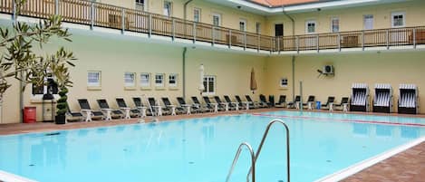 Une piscine dotée de chaises longues et d’un espace salon couvert, le tout entouré d’un bâtiment avec un toit en tuiles et un balcon.