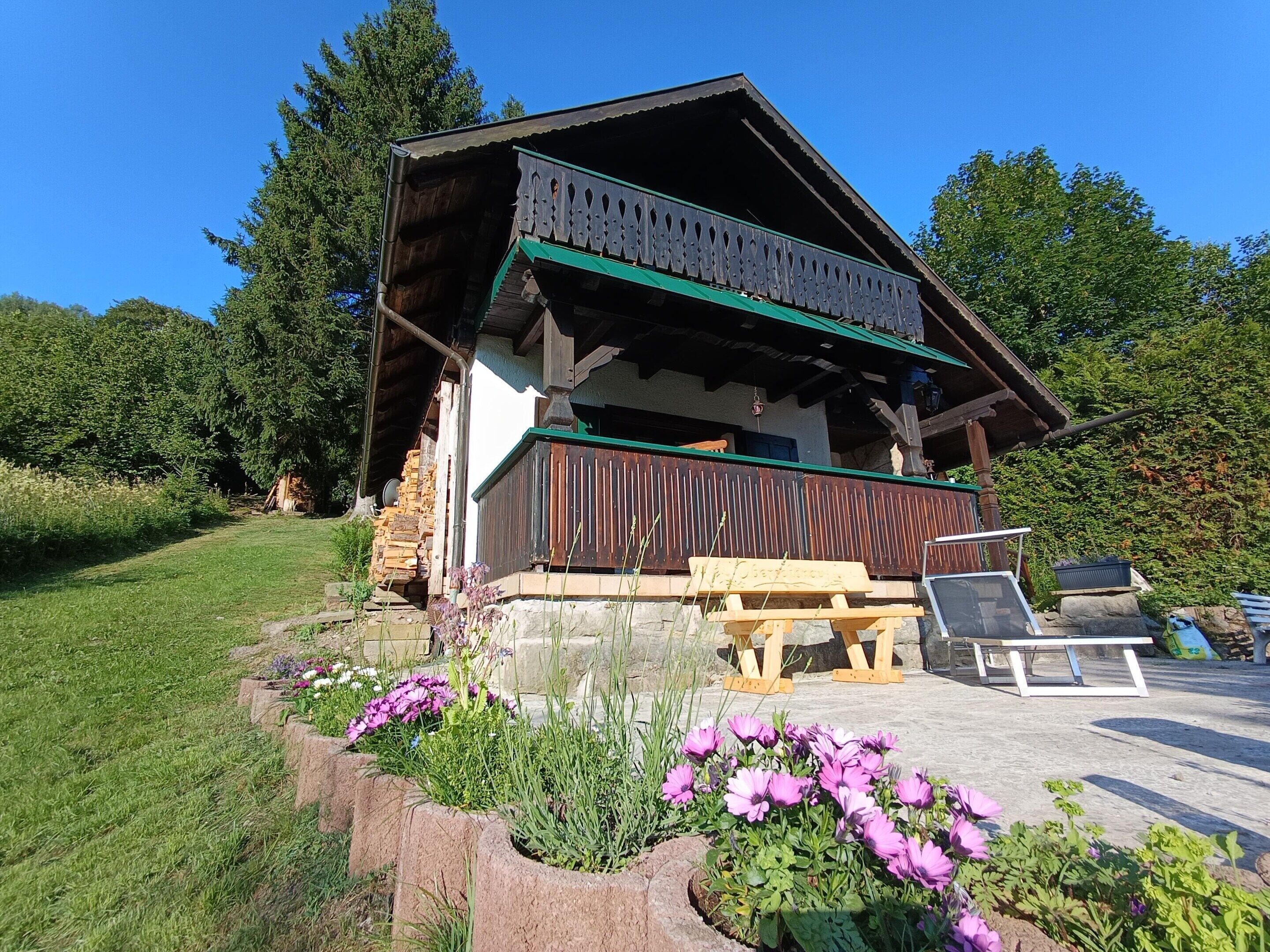 A wooden chalet with a balcony, surrounded by a garden with purple flowers.
