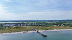 A coastal area with a pier extending into the water, a beach with sun loungers, and a backdrop of wind turbines and industrial buildings. - Ferien- und Freizeitpark Weissenhauser Strand (Wangels)