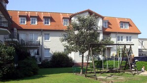A residential building with a red-tiled roof, multiple windows, and a small garden area with a swing set. - Appartementhaus Jann-berghaus, Norderney (Norderney)