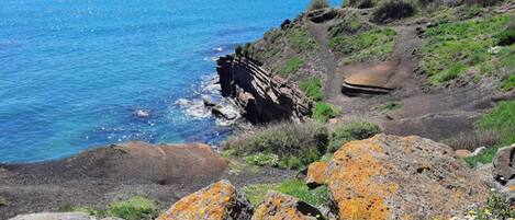 A coastal landscape with rocky terrain, sparse vegetation, and a clear blue sea.