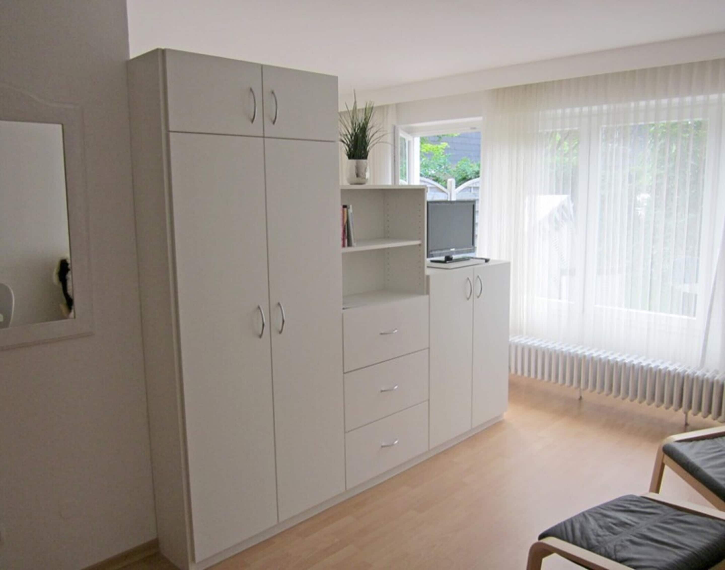 Huoneessa on suuri vaatekaappi, pieni työpöytä, jossa on tietokone, tuoli ja ikkuna, jossa on verhot.