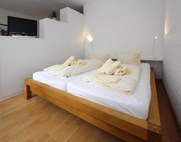 A modern bedroom with a wooden bed, white linens, and a flat-screen TV on a white wall.