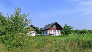 Two traditional half-timbered houses with a sloped roof, surrounded by lush greenery and a clear blue sky.