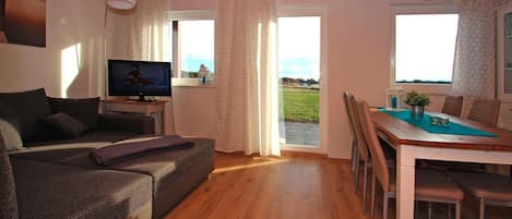 A living room with a sofa, TV, dining table, chairs, and large windows with curtains.