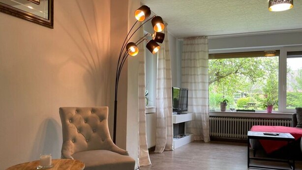 A living room with a tufted armchair, a small round table, a floor lamp, a television, and a view of greenery outside. - Ferienhaus, Friederikensiel (Wangerland)