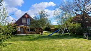A house with a sloped roof, a front porch, and a backyard with a grassy lawn and a blue slide. - Ferienhaus, Friederikensiel (Wangerland)