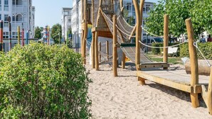 En legestruktur i træ med en rebsbro og en rutsjebane, omgivet af sand og grønne planter, med bygninger i baggrunden.