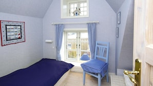 A bedroom with a single bed, a chair, a window with curtains, a small table, and a wall-mounted picture.