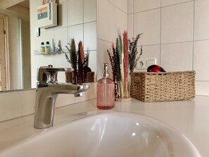 A bathroom with a white sink, a chrome faucet, and a mirror. There are decorative plants and a wicker basket on the counter. - Holiday Home Seerose With Sauna on Lake Dummer, Dummer (Dümmer)