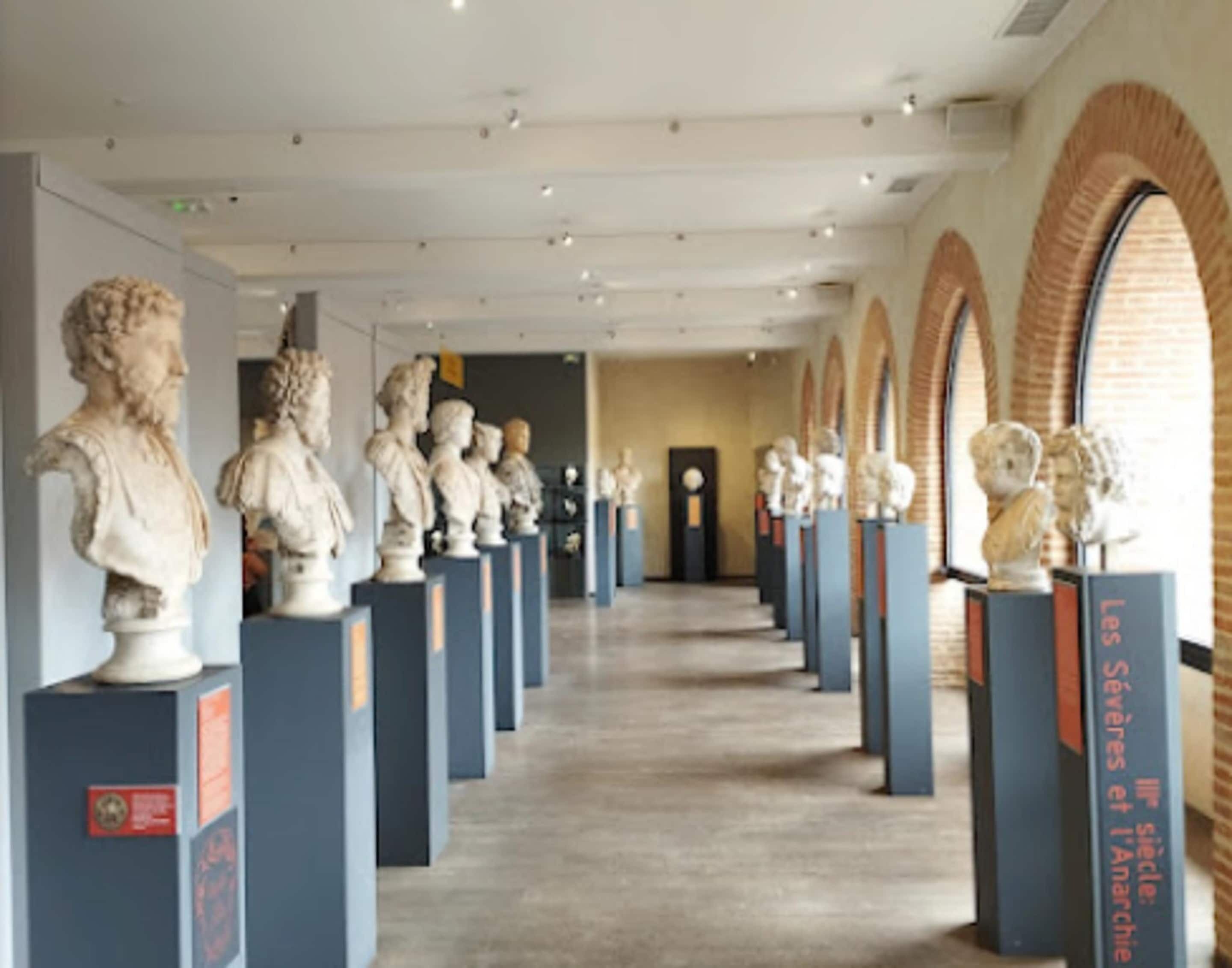 A museum corridor with classical sculptures on pedestals, a brick wall on one side, and a door at the end.