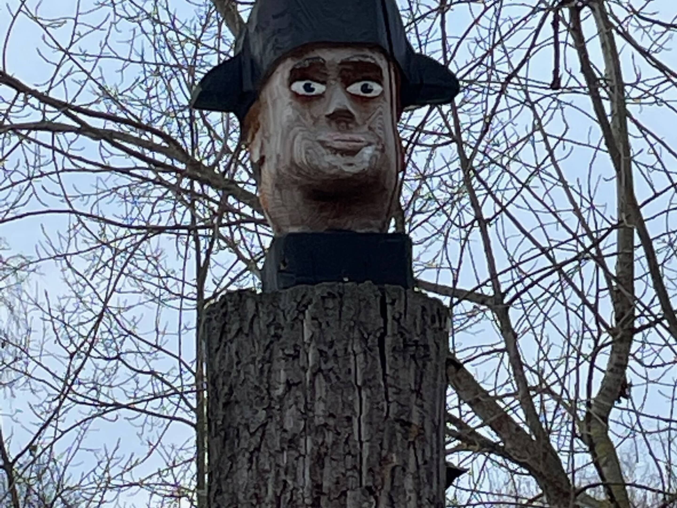 Une sculpture en bois représentant un personnage coiffé d’un chapeau, posée sur le tronc d’un arbre.