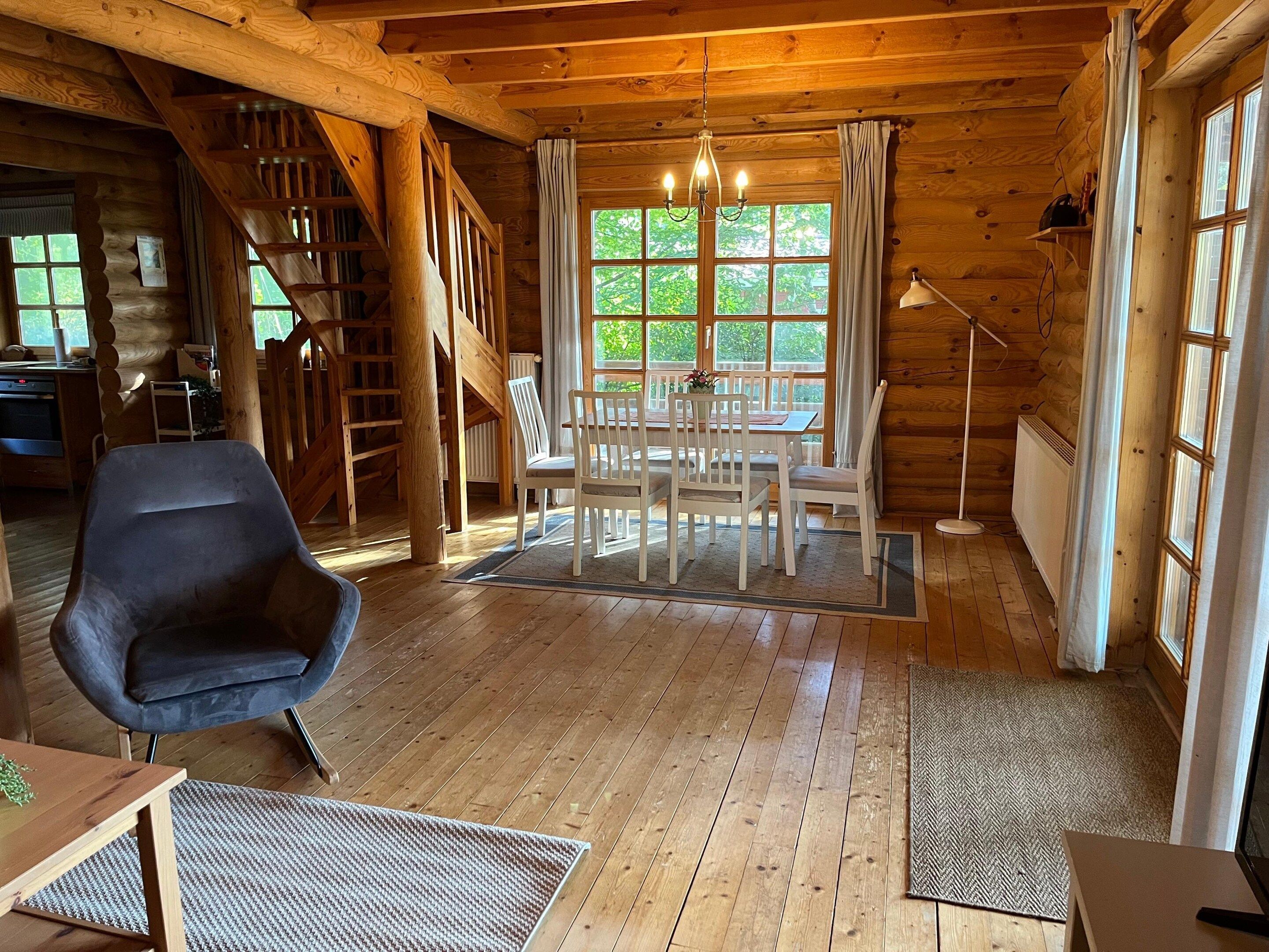 Interior de uma cabana de madeira com área de refeições, escada e vista através de grandes janelas.