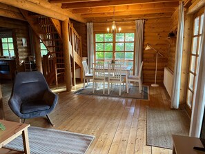 Interior de uma cabana de madeira com área de refeições, escada e vista através de grandes janelas.