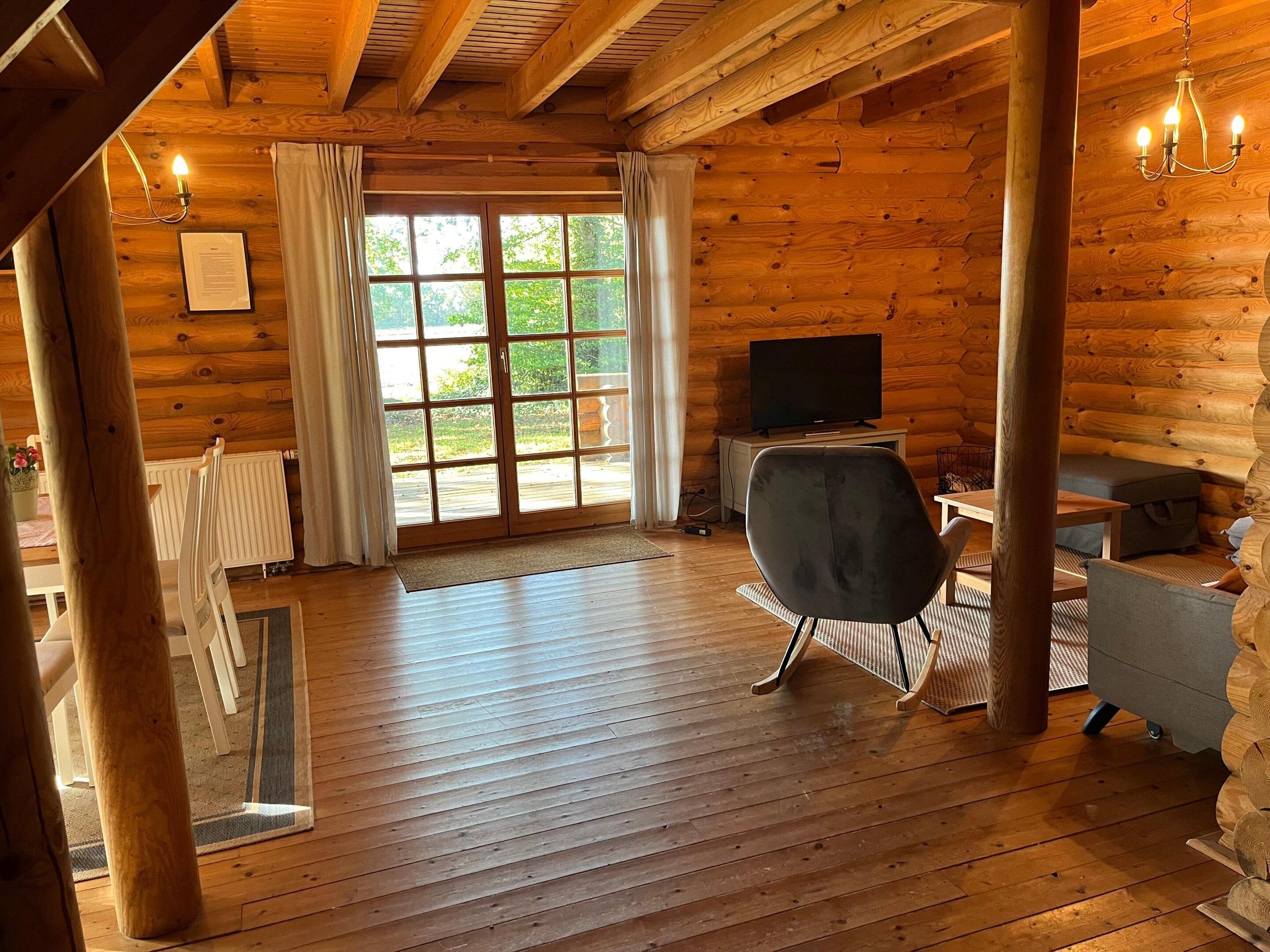 Interior de uma cabana rústica com piso de madeira, uma televisão sobre um suporte, uma cadeira preta e uma janela com cortinas.