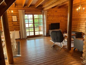 Interior de uma cabana rústica com piso de madeira, uma televisão sobre um suporte, uma cadeira preta e uma janela com cortinas.