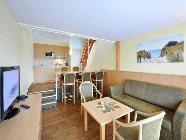 Ett kompakt bostadsutrymme med ett litet kök, en matplats med stolar, en soffa, en tv och en tavla som föreställer en strandscen.