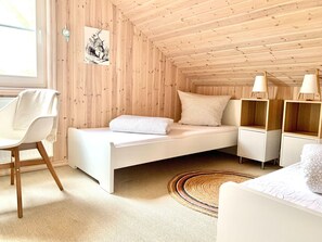 A cozy room with a wooden sloped ceiling, a white bed with a pillow, a white chair with a towel, and a small wooden shelf with a lamp. - Nice Holiday Home in Dummer Near Lake (Dümmer)