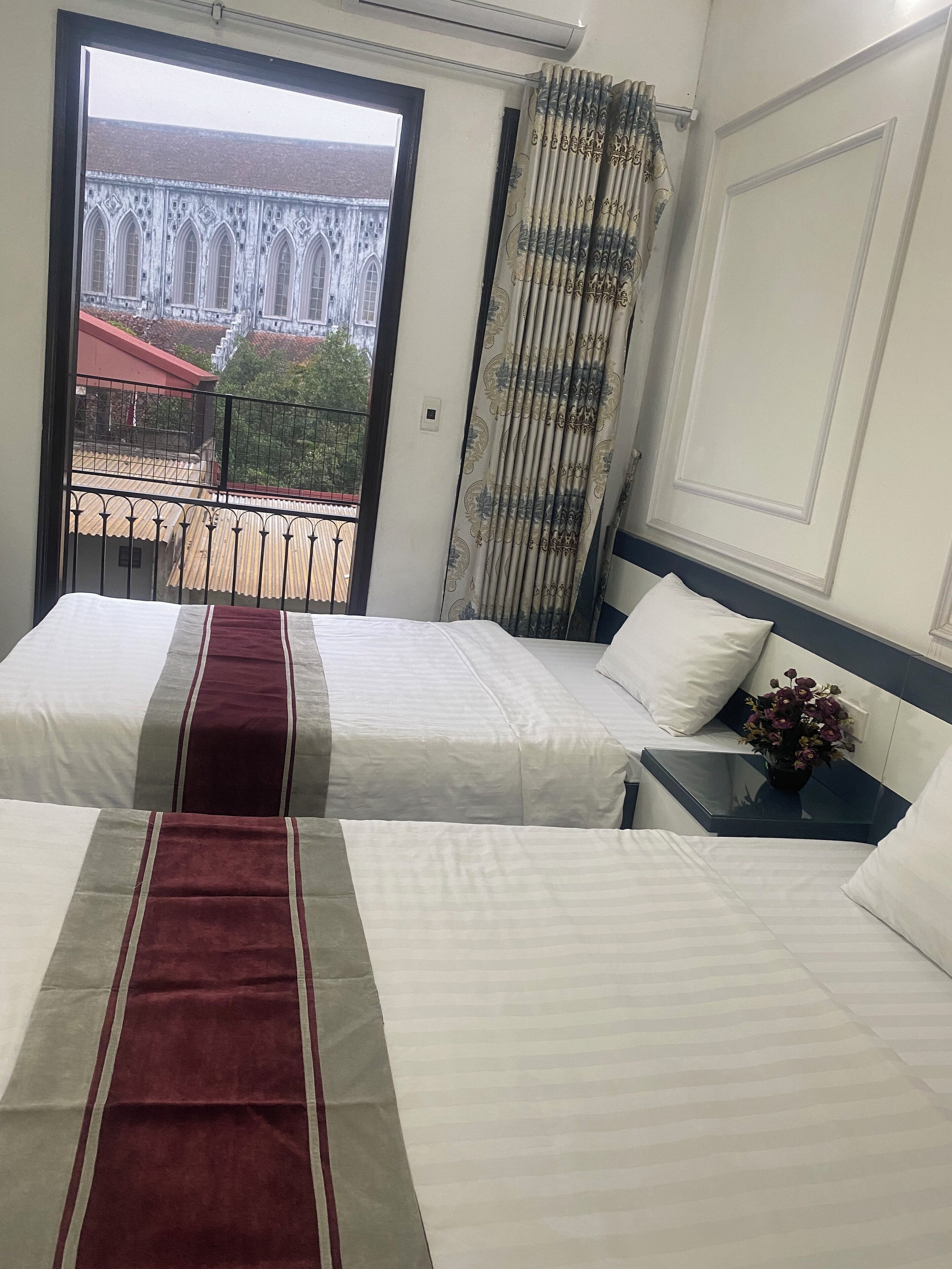Une chambre d’hôtel avec deux lits, une table de chevet ornée d’un vase de fleurs et une vue sur un bâtiment, visible à travers une porte ouverte.