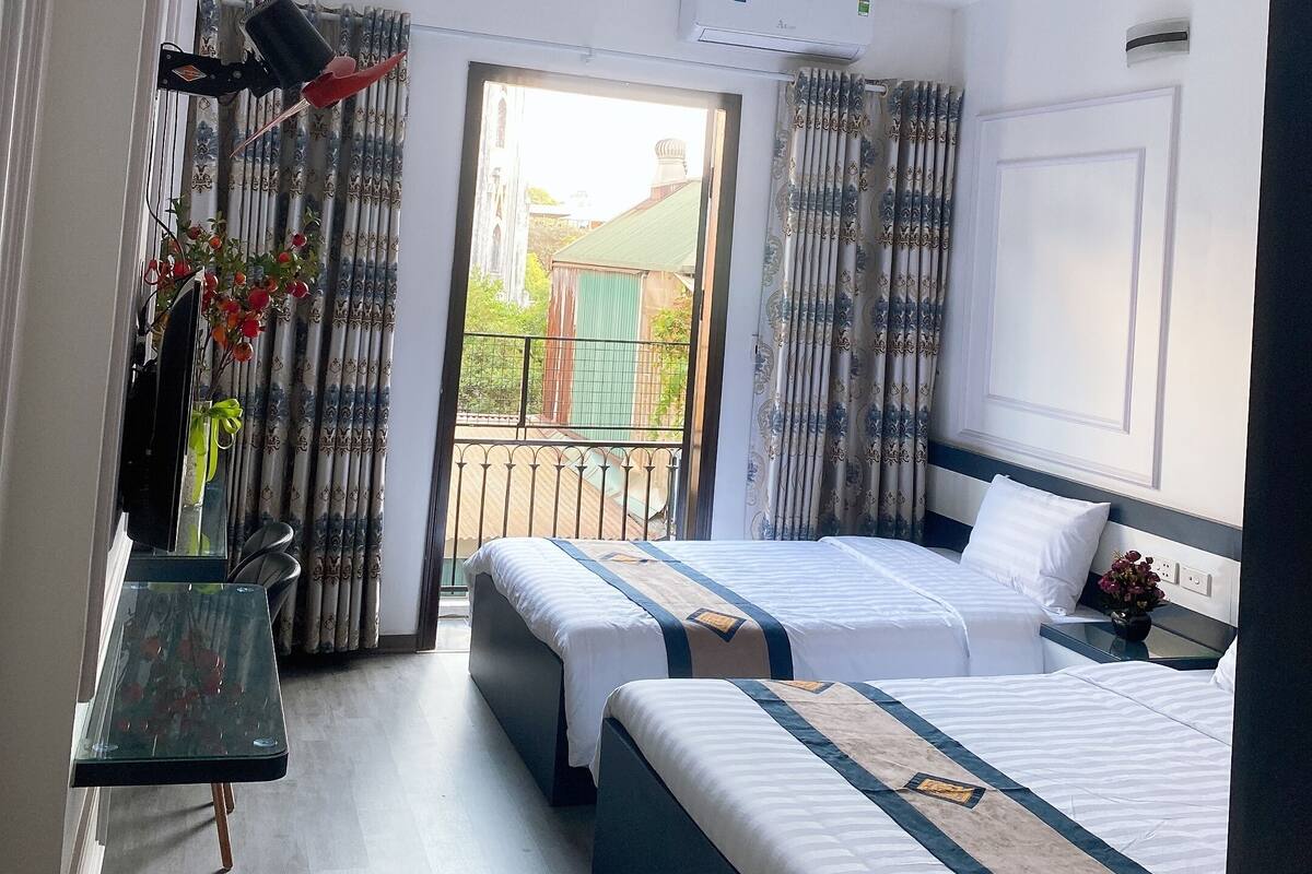 A hotel room with two beds, a desk with a chair, an air conditioning unit, and a view of the outside.