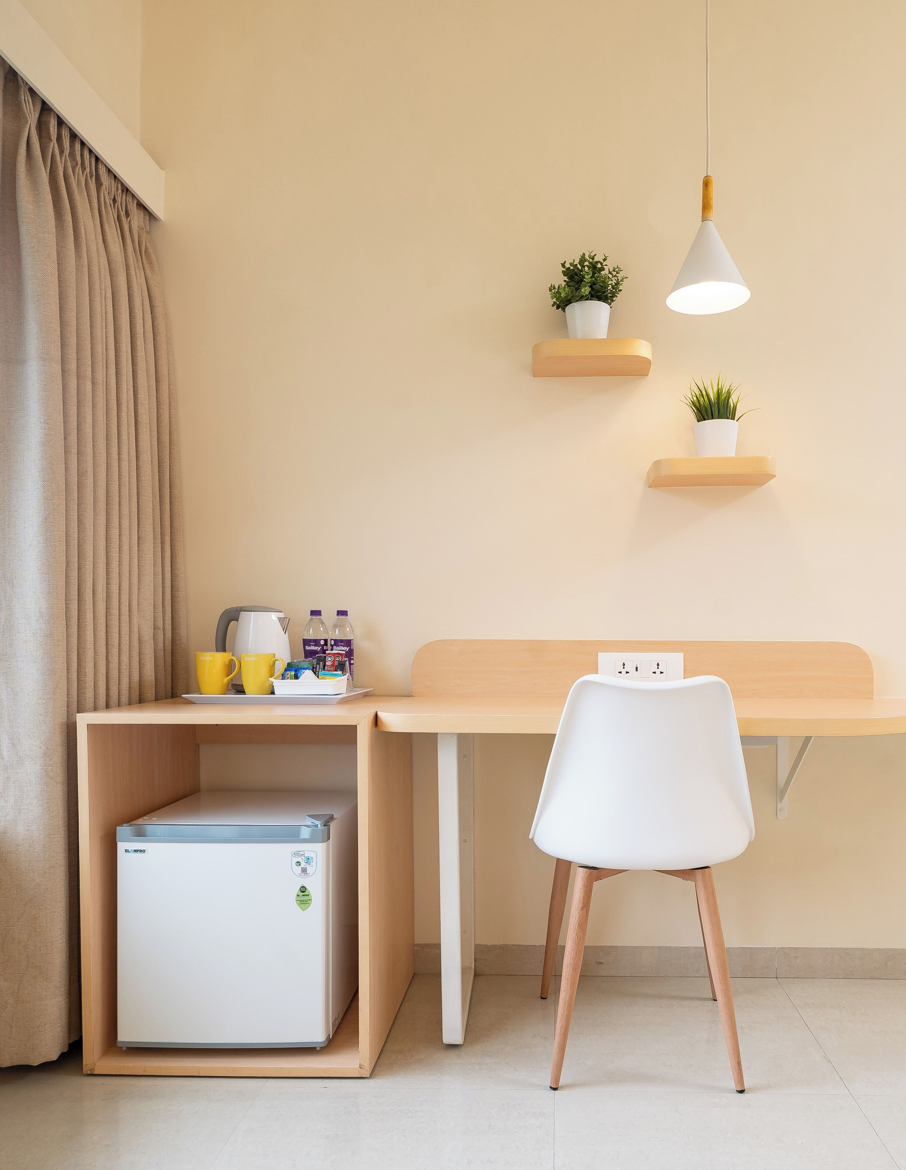 Habitación pequeña con escritorio de madera, una silla blanca, un mini refrigerador, una tetera y dos plantas en estantes fijos a la pared.