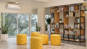A modern library with large windows, a bookshelf, and yellow ottomans. - Bloom Hotel Khandala (Mawal)